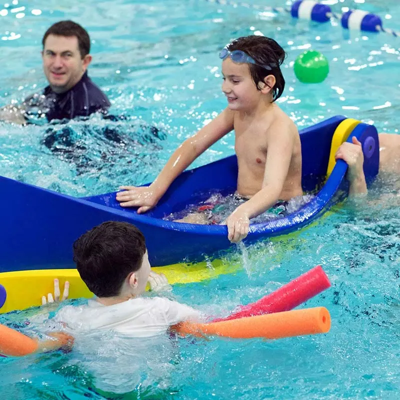 Children in a pool with a coach, using a large float and foam noodles for support