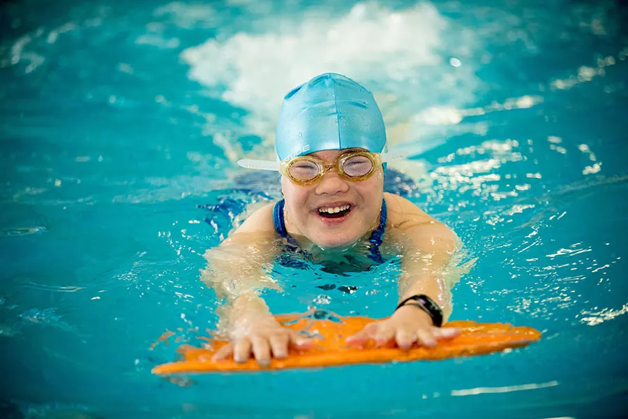 A smiling swimmer in a blue cap and yellow goggles kicking across the pool on an orange kickboard