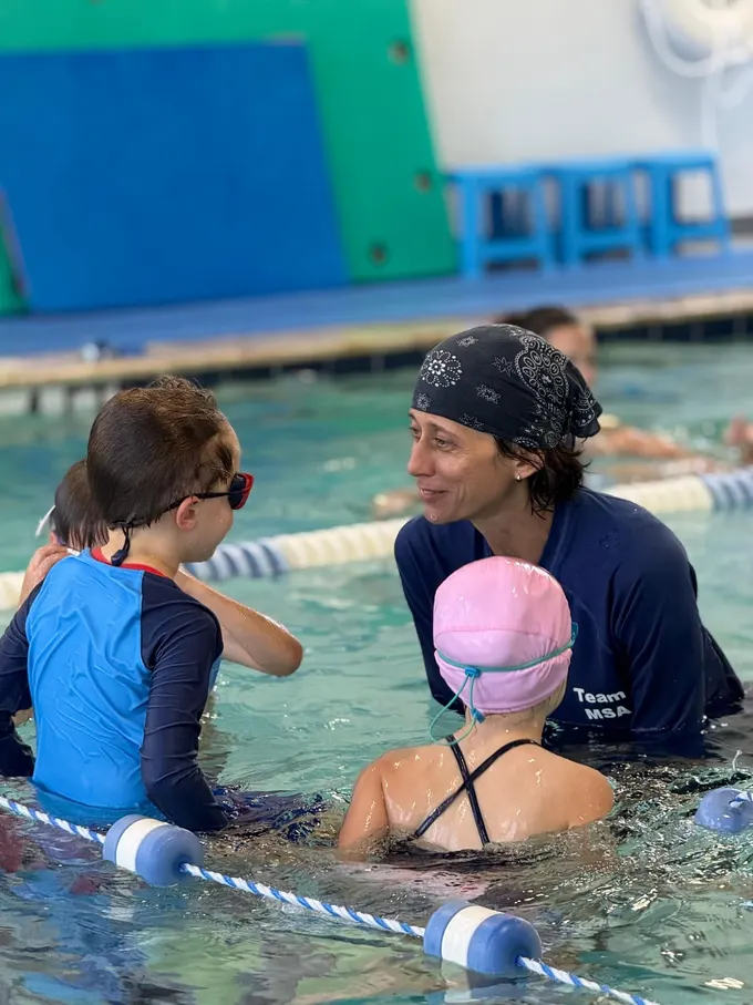 A coach in a Team MSA rash guard at the lane rope with two children, one in a blue rash guard and one in a pink cap