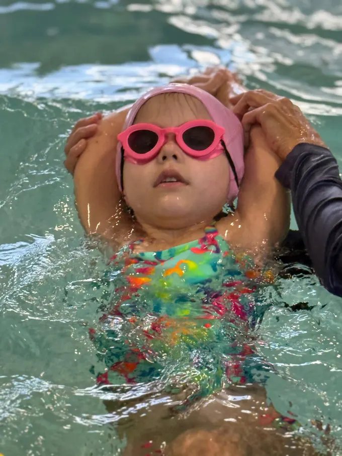 A small child in a pink cap and pink goggles floating on her back, held by an instructor