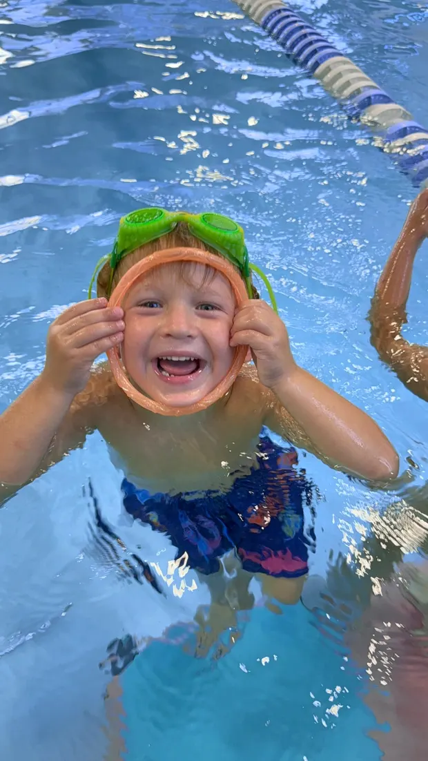 A boy laughing in the pool, green goggles pushed up on his head and an orange mask ring held around his face