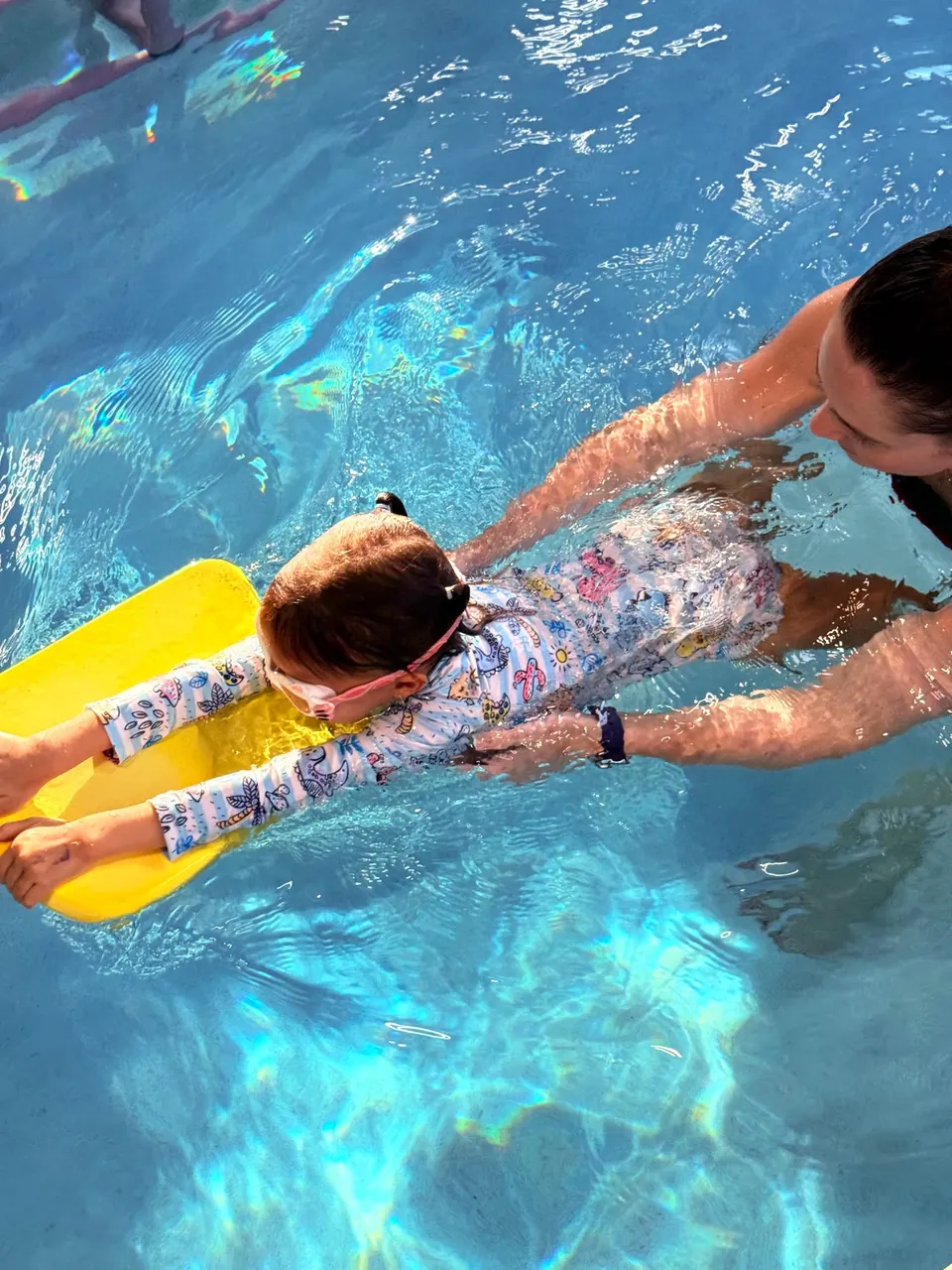 Seen from above: a young girl in a patterned long-sleeved swimsuit and pink goggles gliding across the pool on a yellow kickboard, an instructor supporting her from behind
