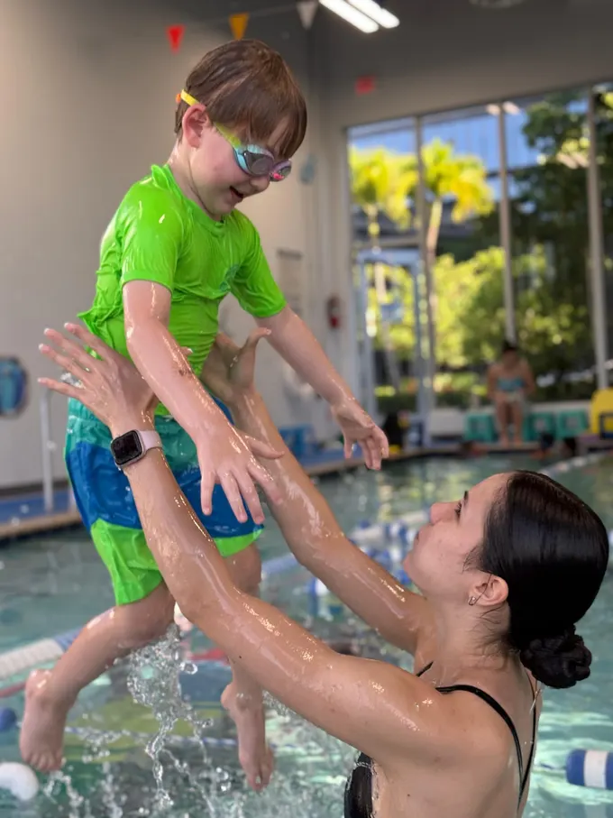 An instructor lifting a laughing boy in a green rash guard clear out of the water