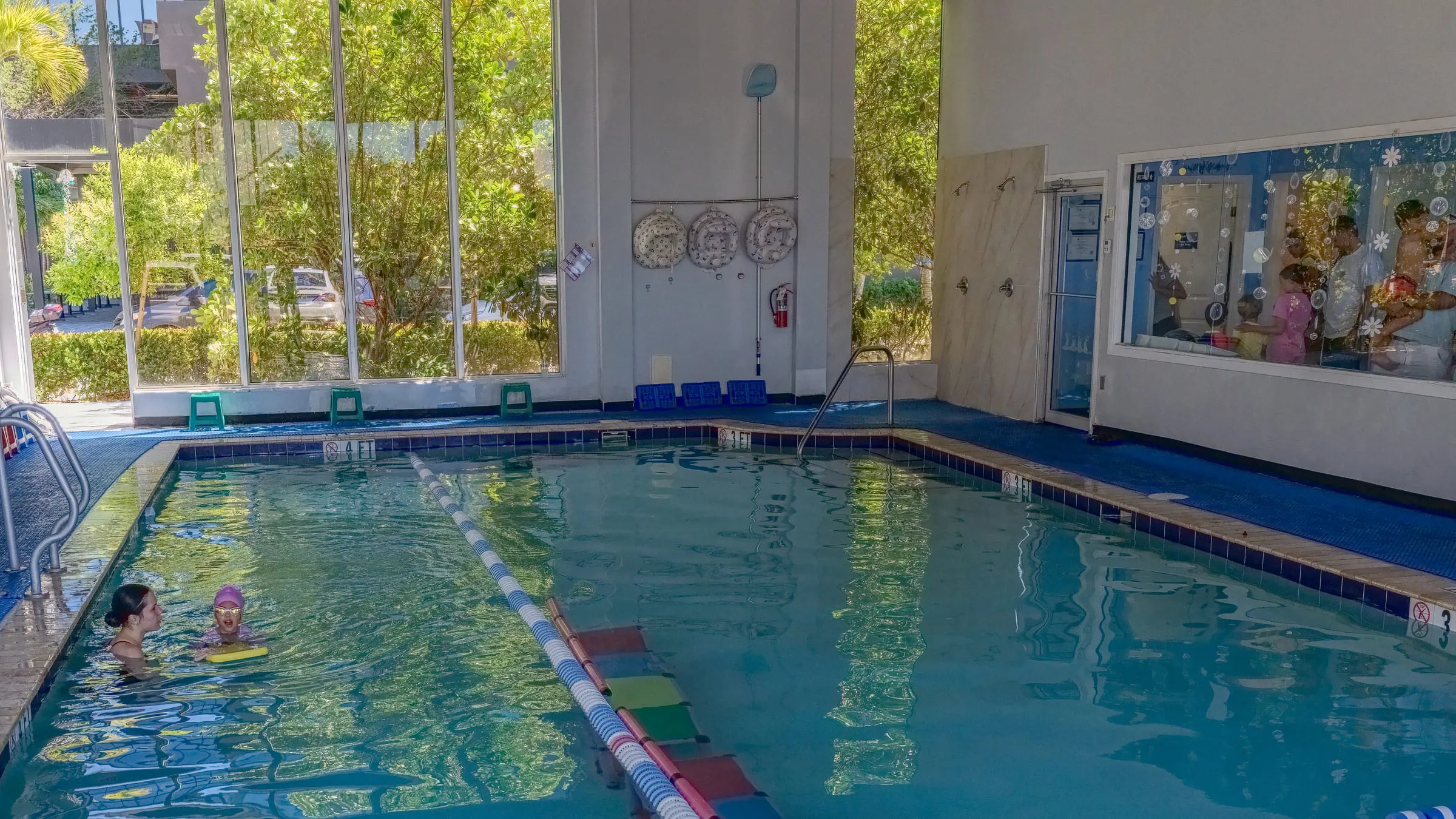 The indoor pool from the deck: a two-storey wall of glass onto tropical greenery, coloured stools along the tiled edge, a lane rope down the middle, and an instructor steadying a kickboard for a child in the shallow end
