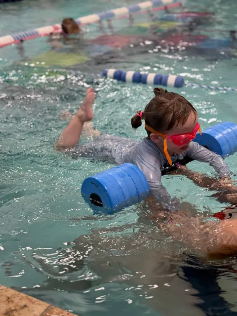 A girl in goggles kicking across the pool with a blue foam bar held out in front of her