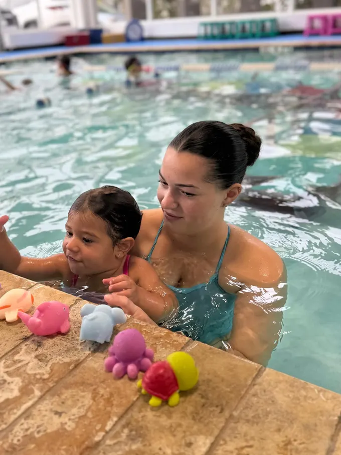An instructor holding a toddler at the pool edge, reaching for bath toys on the tiles