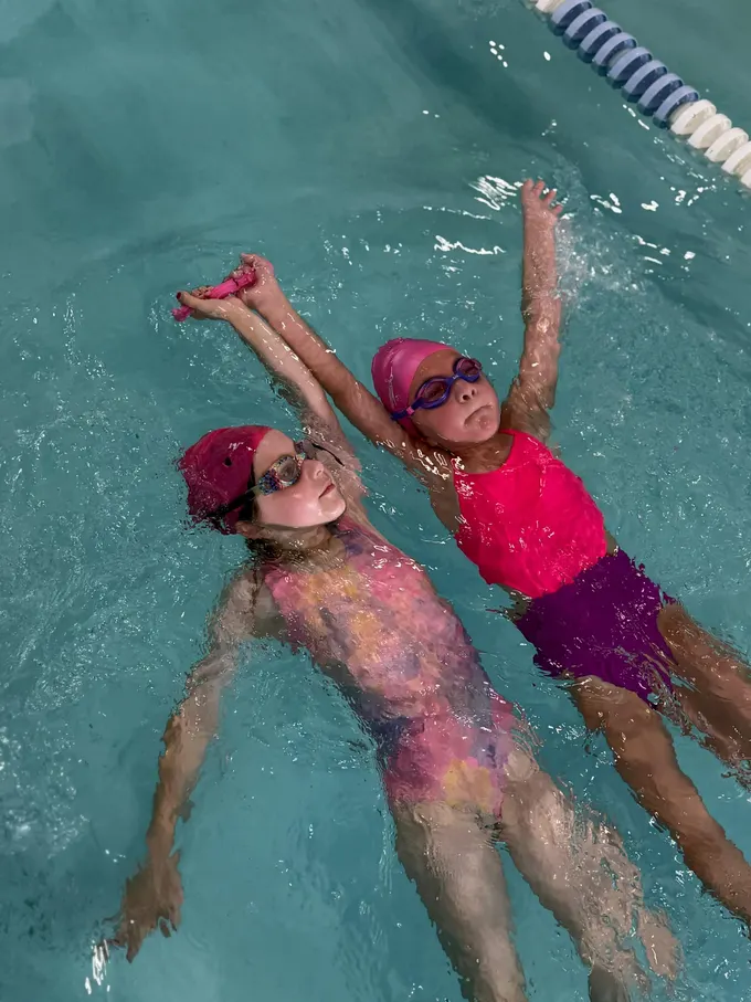 Two girls in pink caps gliding side by side on their backs, arms stretched overhead